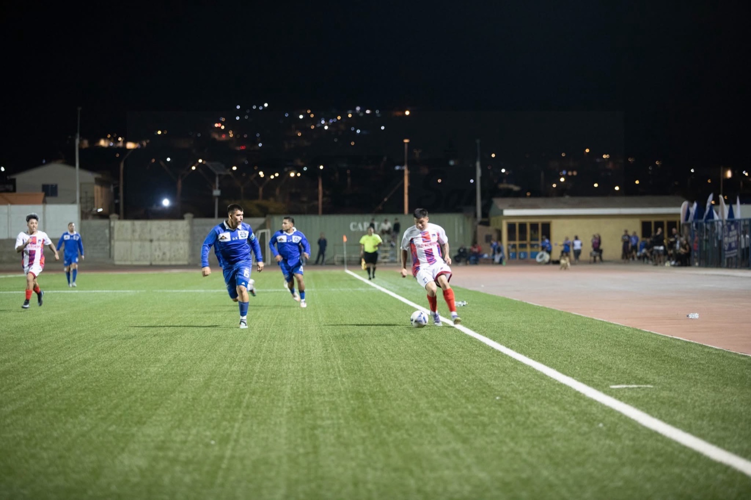 Acción por la banda con control de balón y persecución de jugadores, en partido nocturno de San Antonio Norte de Taltal.