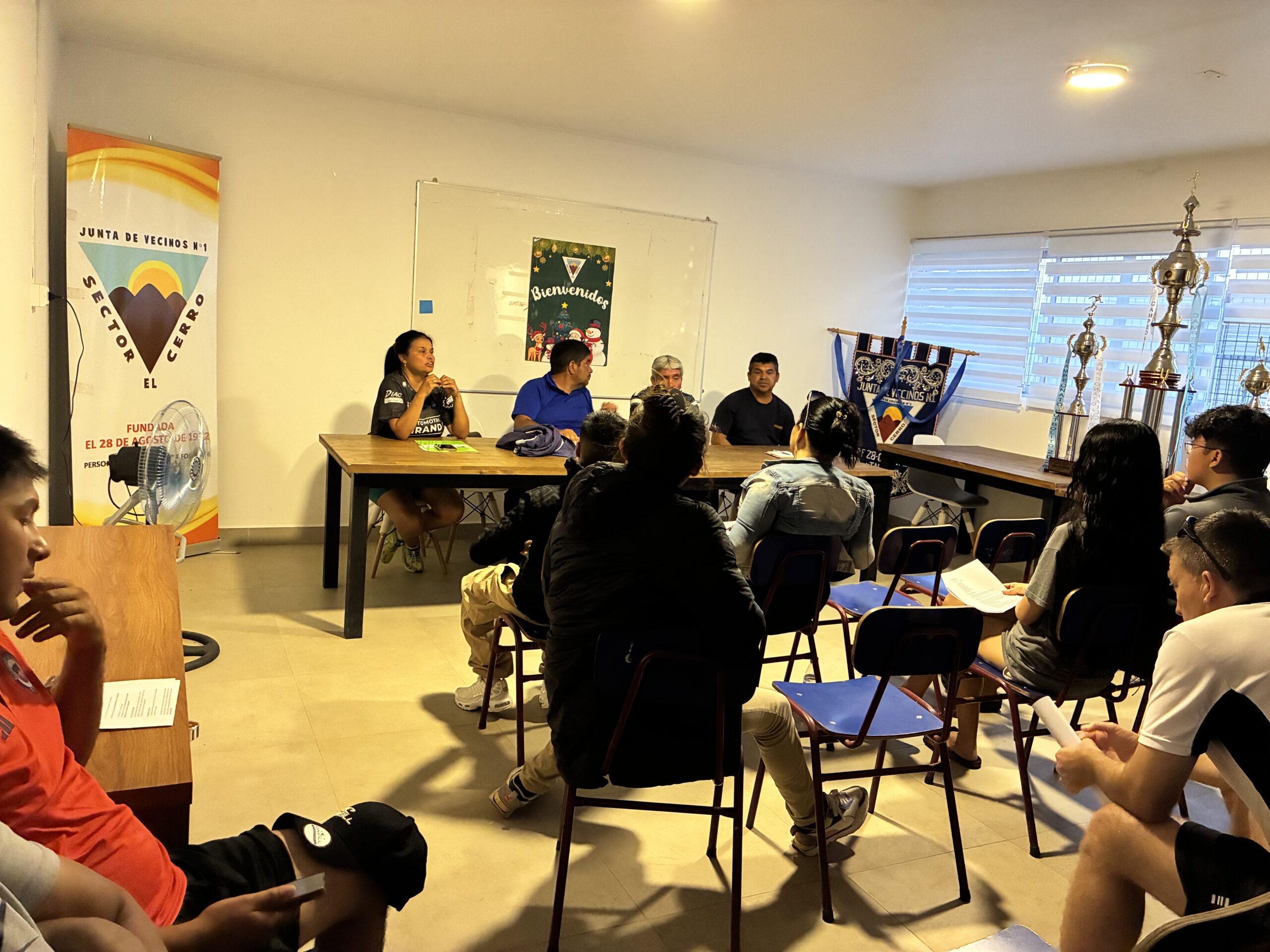 Reunión informativa en la Junta de Vecinos Número 1 Sector El Cerro con directiva y asistentes junto a un pendón institucional.