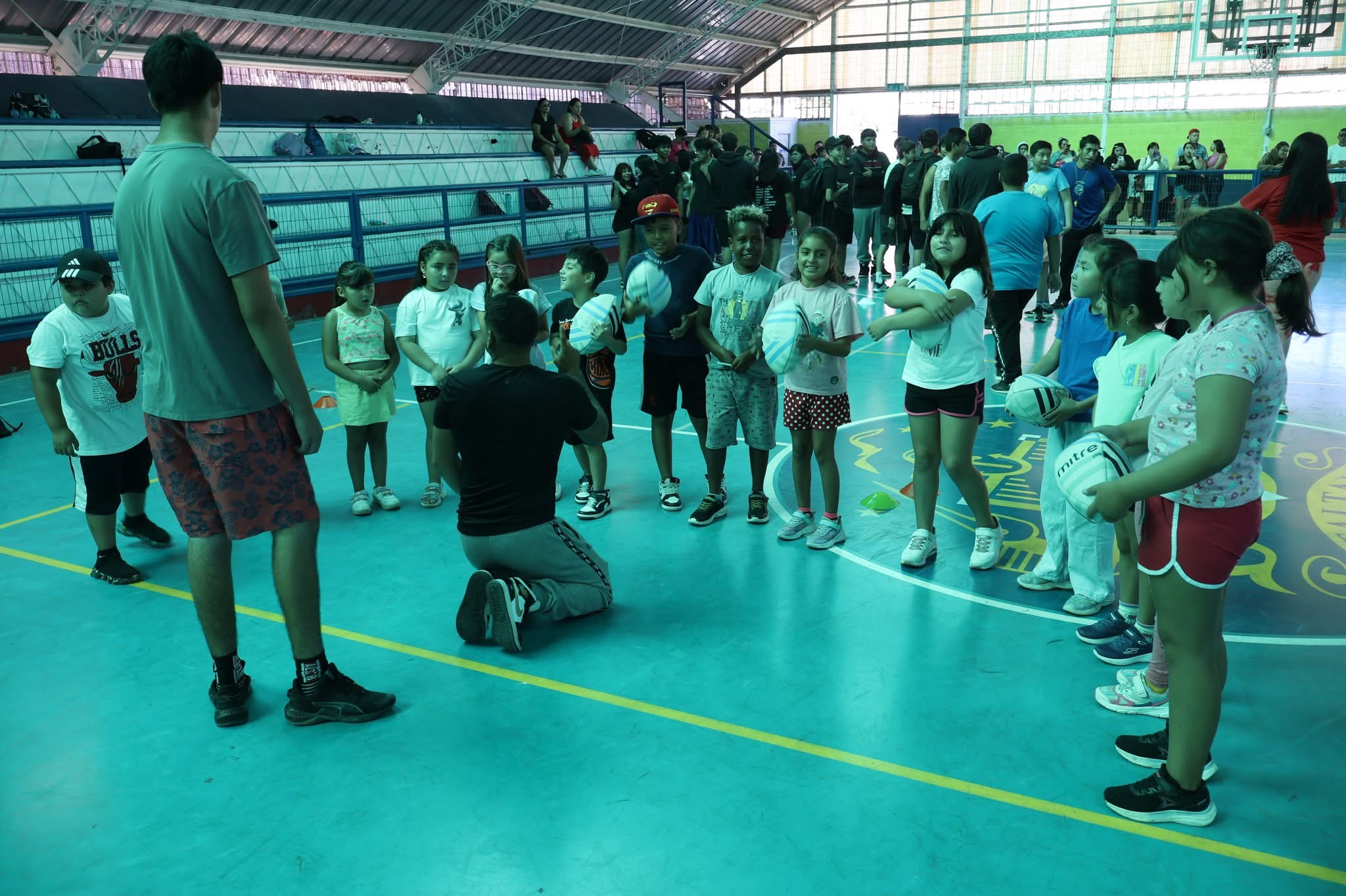 Niños pequeños con balones de rugby recibiendo instrucciones de un monitor durante la jornada de iniciación deportiva.