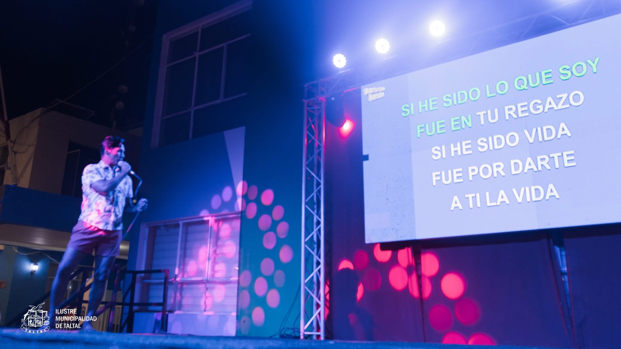 Escenario municipal con pantalla gigante proyectando letras de canciones para el público.