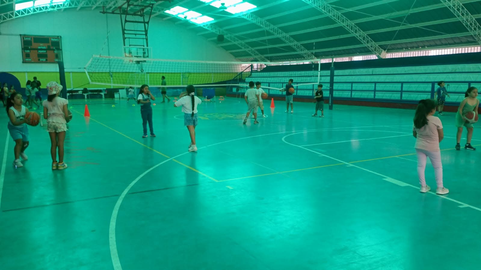 Grupo de niñas practicando fundamentos de básquetbol en gimnasio techado.