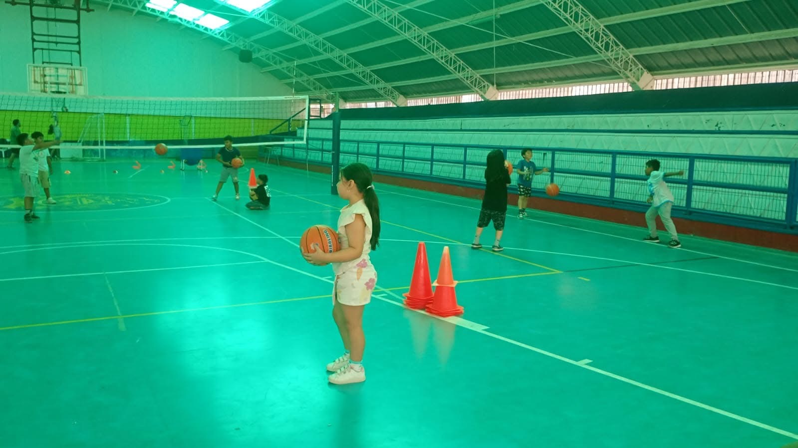 Niña con balón de básquetbol frente a conos en clase de gimnasio.