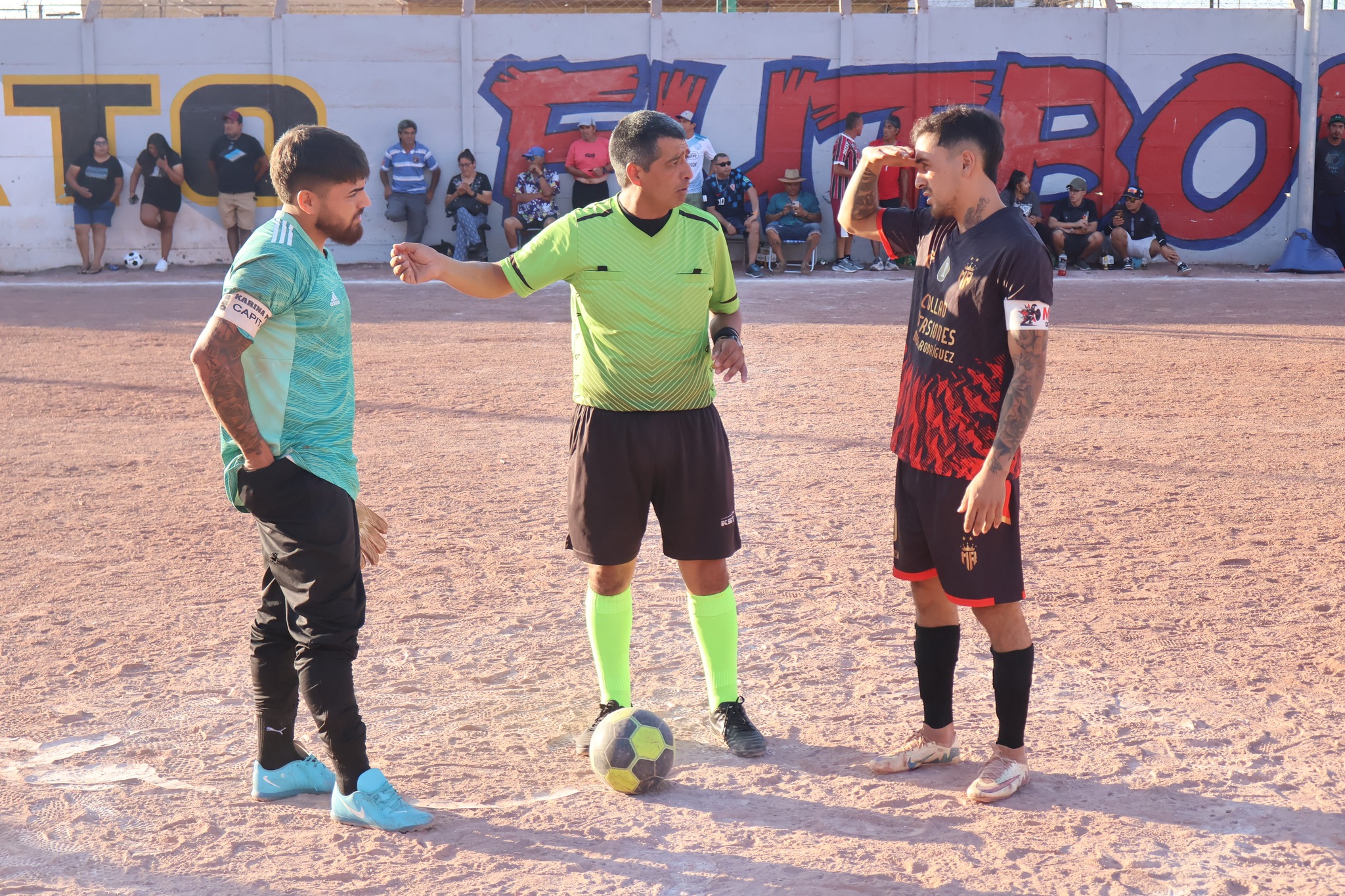Árbitro media discusión entre jugador de Karina FC (brazo extendido) y capitán de Manuel Rodríguez en la cancha durante el Campeonato de Futbolito de Taltal edición 44, febrero 2026. Fondo con muro grafiteado "FUTBOLITO” y público en gradas.