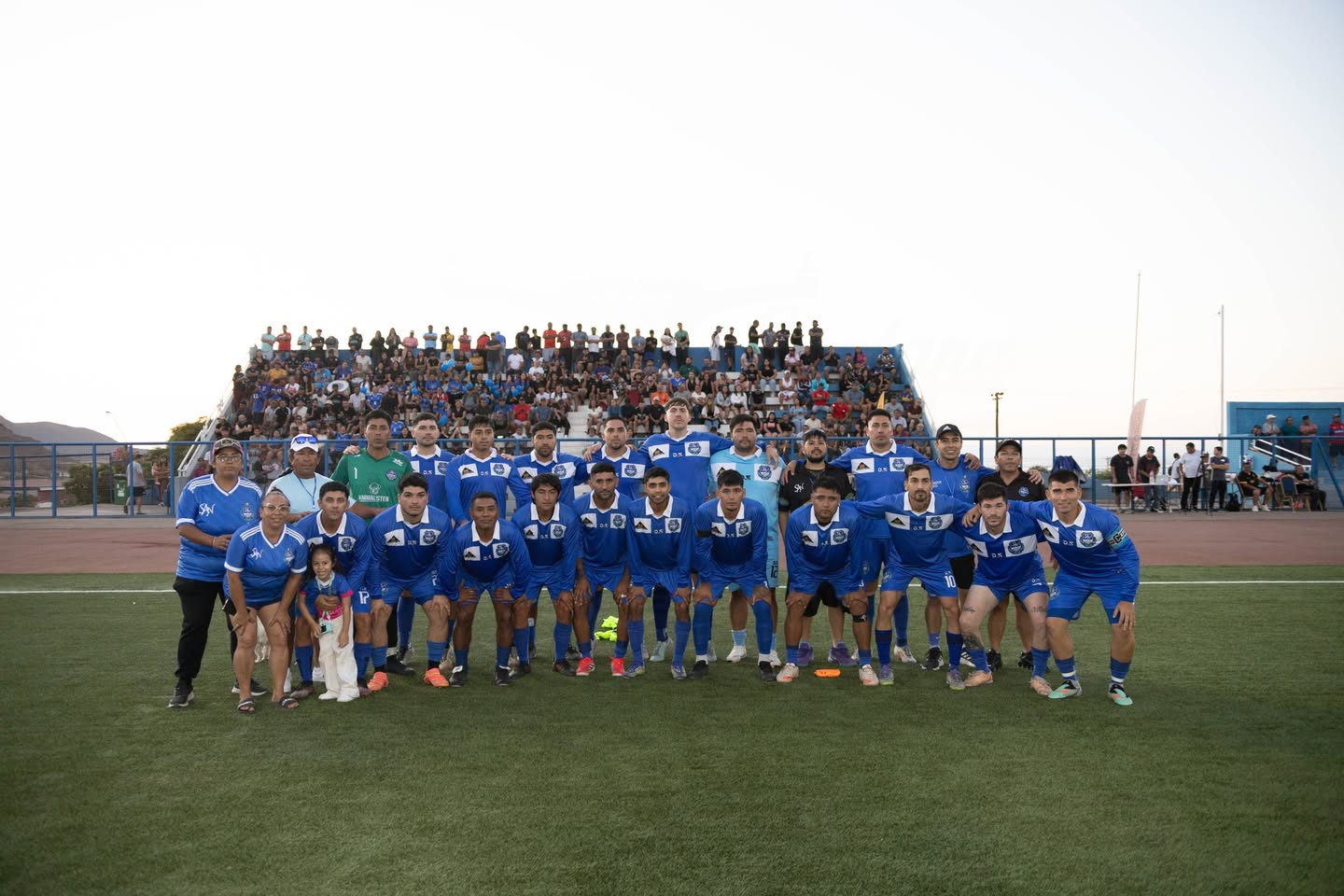 Plantel completo de San Antonio Norte de Taltal posando en el Estadio Belmor Rojas con trofeo y bandera, junto a hinchada en graderío durante el Torneo Zonal Norte ANFA 2026, febrero 2026.