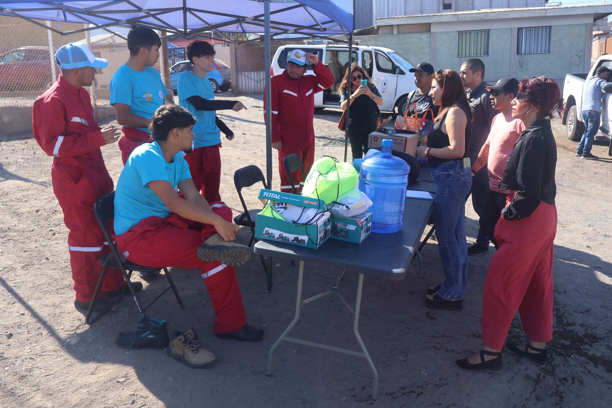 Jóvenes del Team Verano 2026 y equipo municipal conversan bajo carpa en Pasaje Entel, con insumos y donaciones en mesa durante jornada comunitaria de mejoramiento urbano, Taltal, enero 2026.
