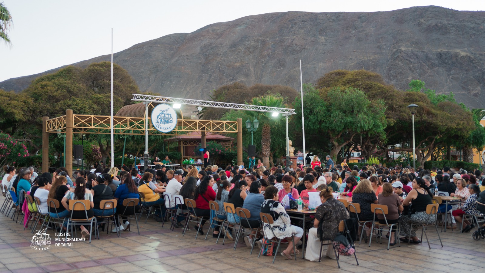 Vista panorámica de cientos de vecinas y vecinos en mesas disfrutando el Gran Bingo Veraniego en Plaza Prat al atardecer, Taltal, Verano 2026.
