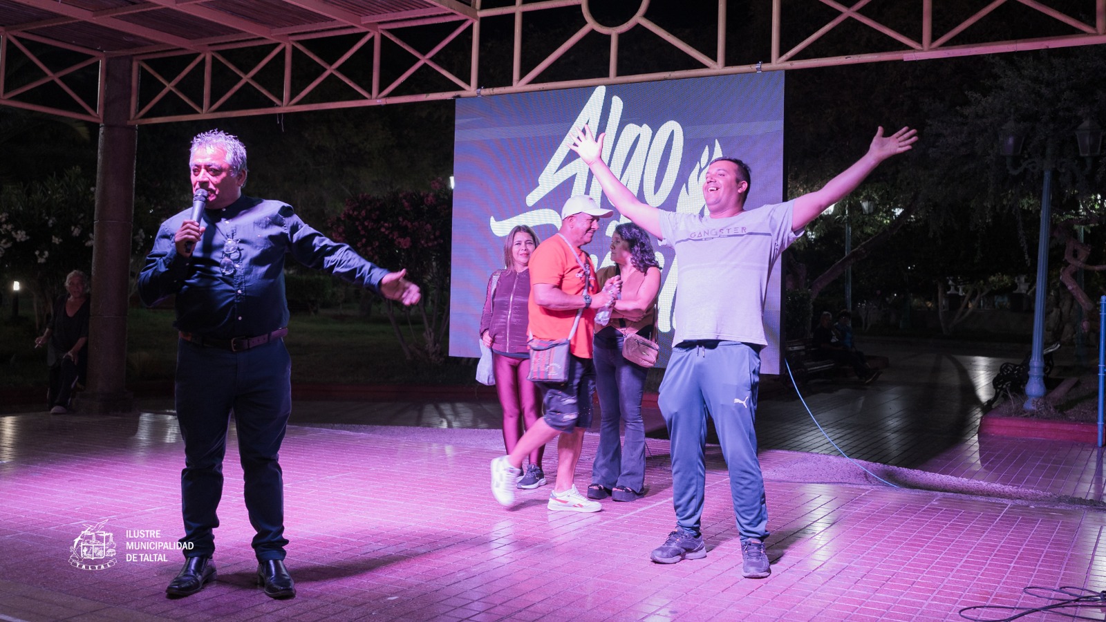 Mario Castillo animando con micrófono el Karaoke de Cumbias en la Plaza de Armas de Taltal, verano 2026. Luces moradas y público disfrutando la noche de música y humor.