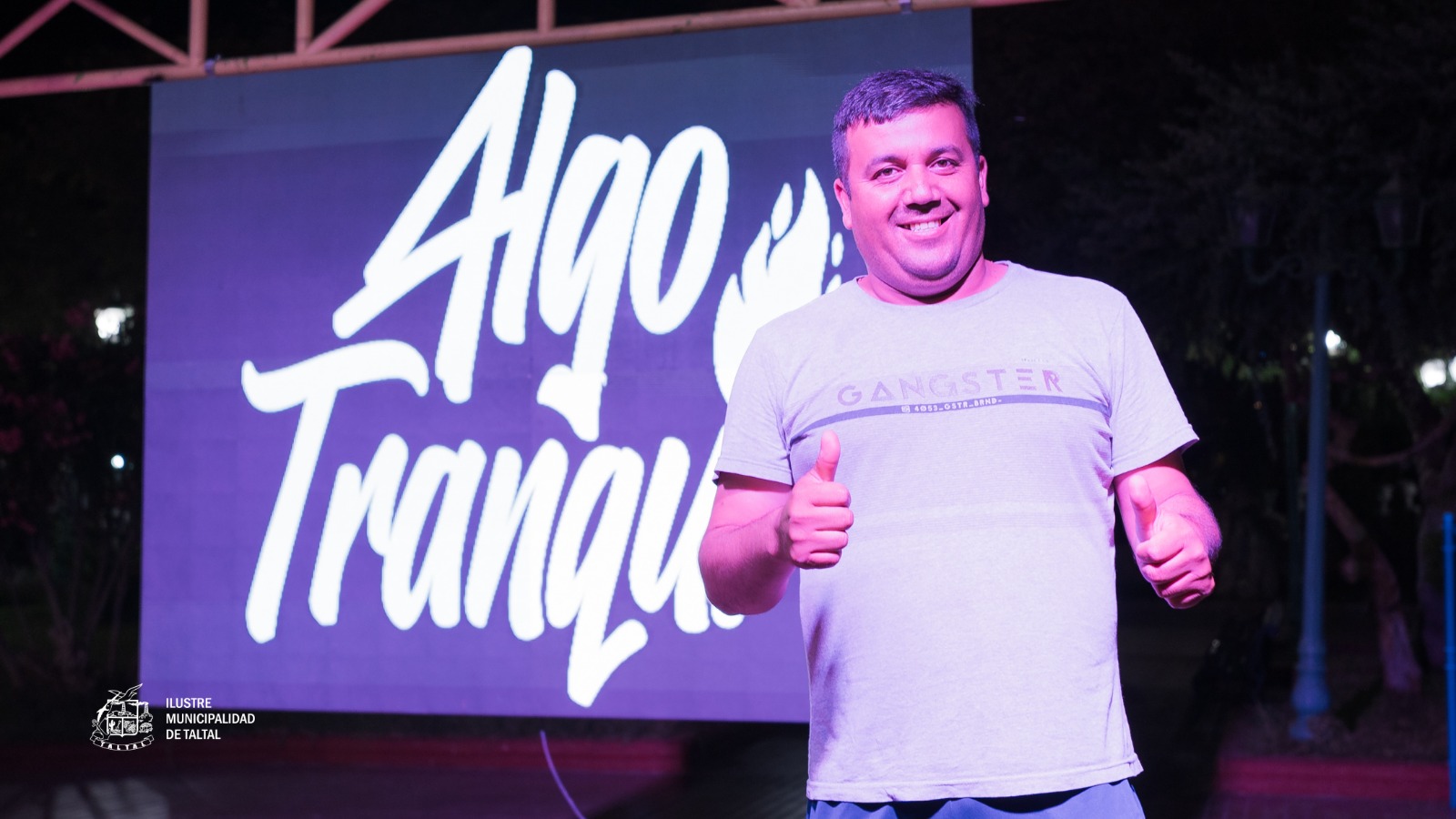 Participante con polera gris y gorra dando thumbs up en el Karaoke de Cumbias organizado por Algo Tranqui en Plaza de Armas de Taltal, febrero 2026.