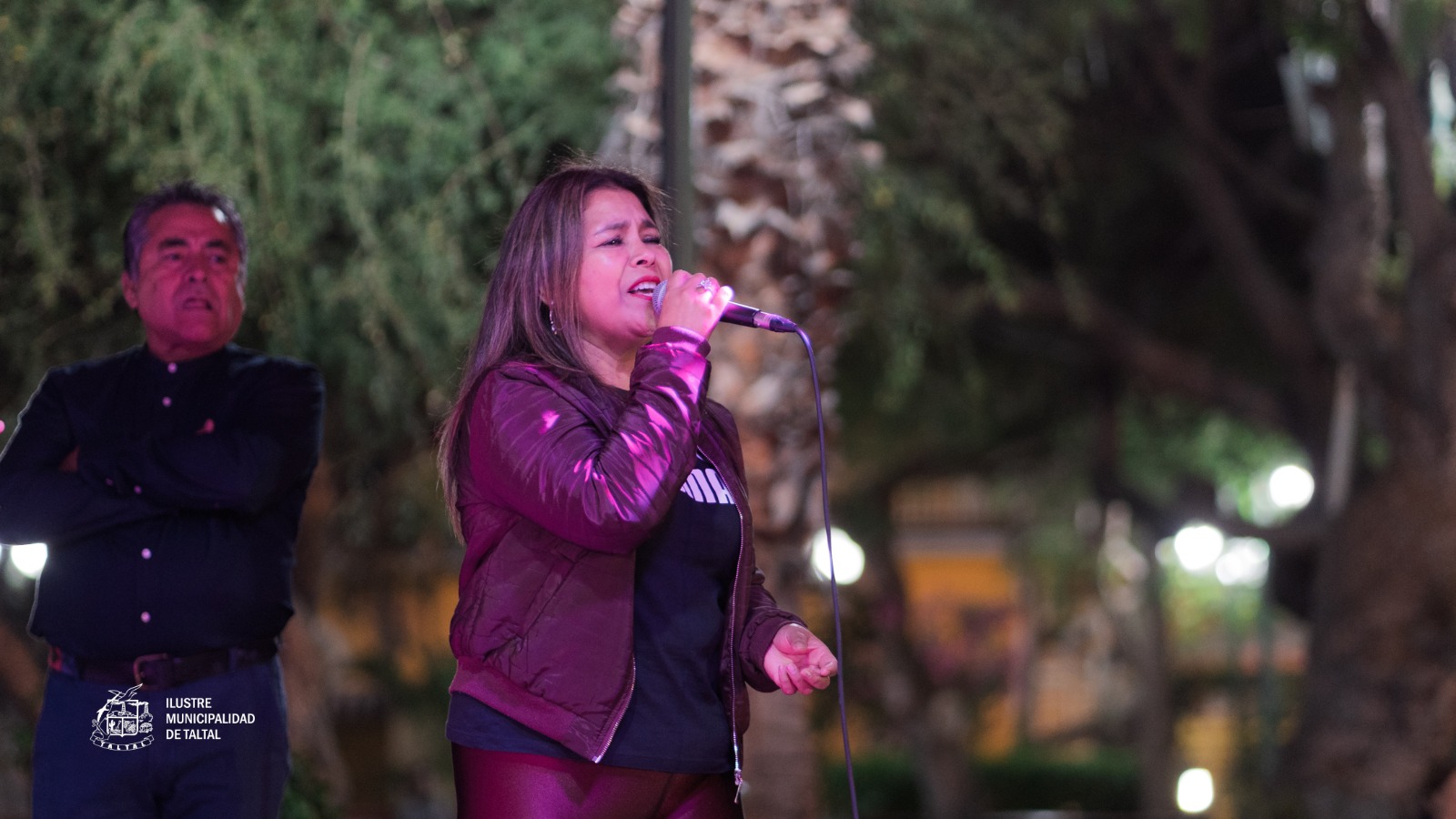 Mujer con chaqueta de cuero cantando en el Karaoke de Cumbias en la Plaza de Armas de Taltal, noche de verano 2026 con luces y ambiente comunitario.