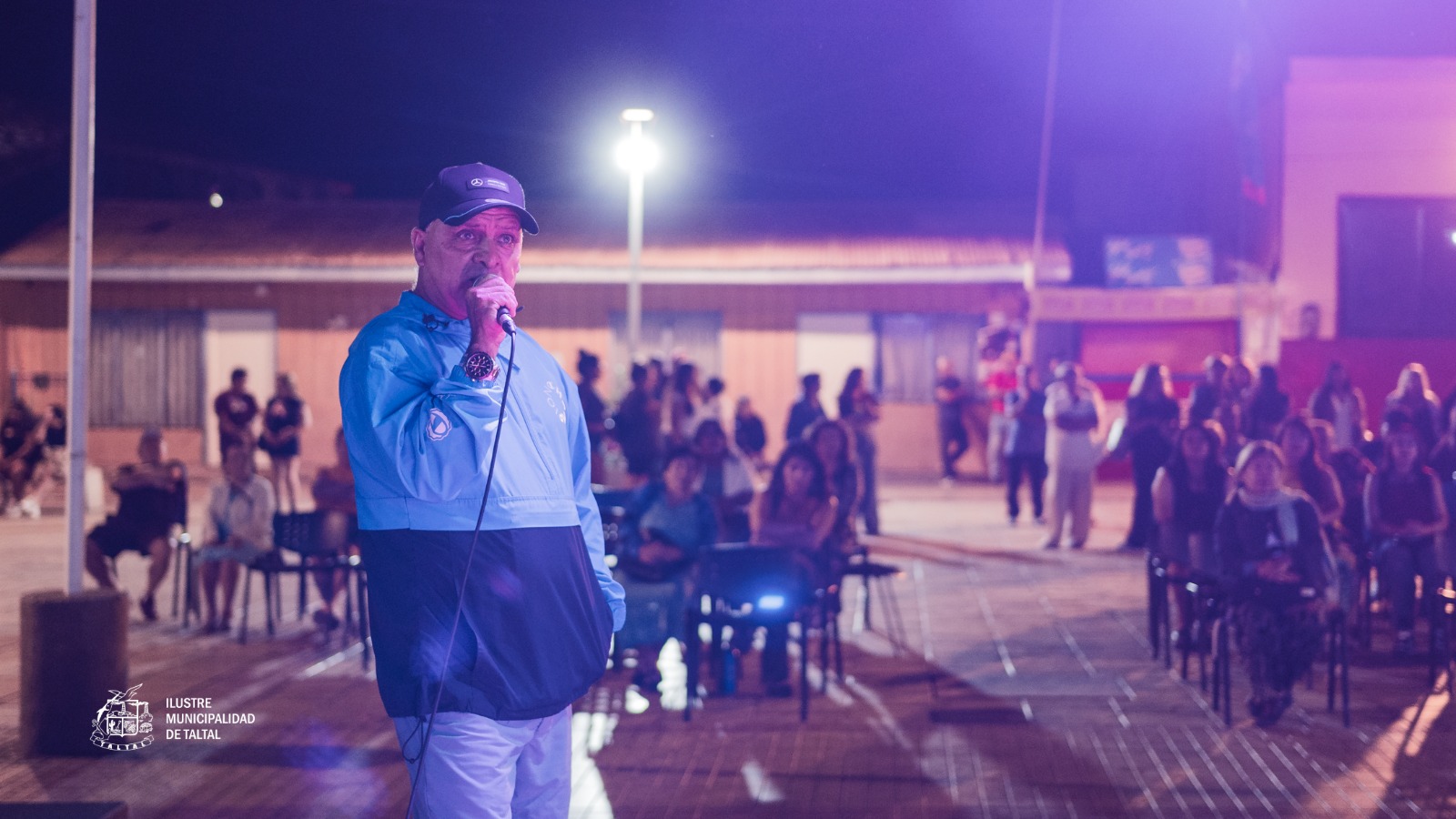 Participante con polera azul cantando con micrófono en el Karaoke de Cumbias en Plaza de Armas de Taltal, noche de verano 2026 con ambiente festivo.