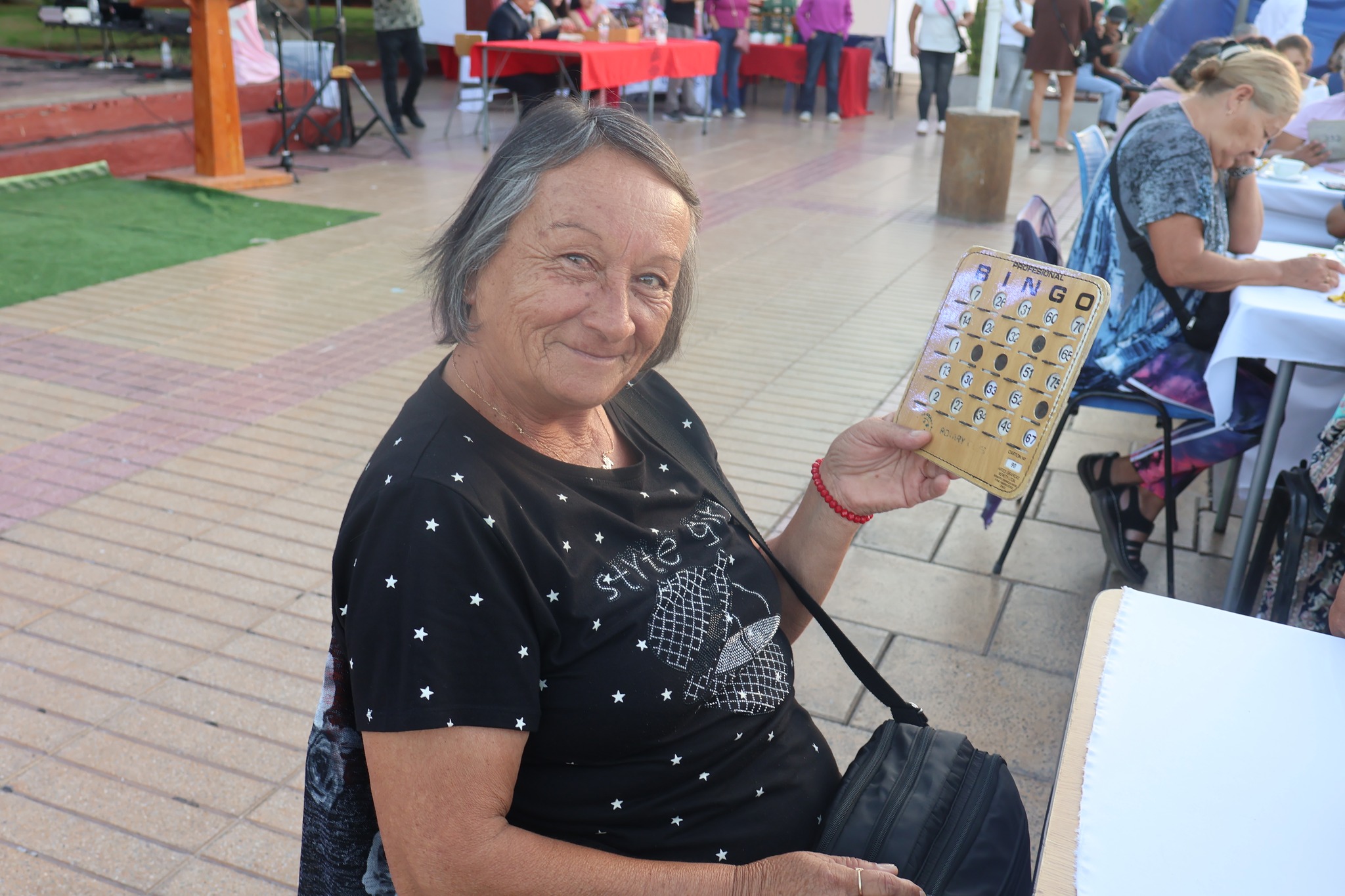 Vecina sostiene cartón de bingo en mesa durante celebración del Día de la Mujer en Plaza de Armas de Taltal.